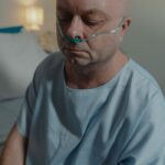 Bald adult man in hospital gown with nasal cannula, sitting indoors, conveying sadness and illness.